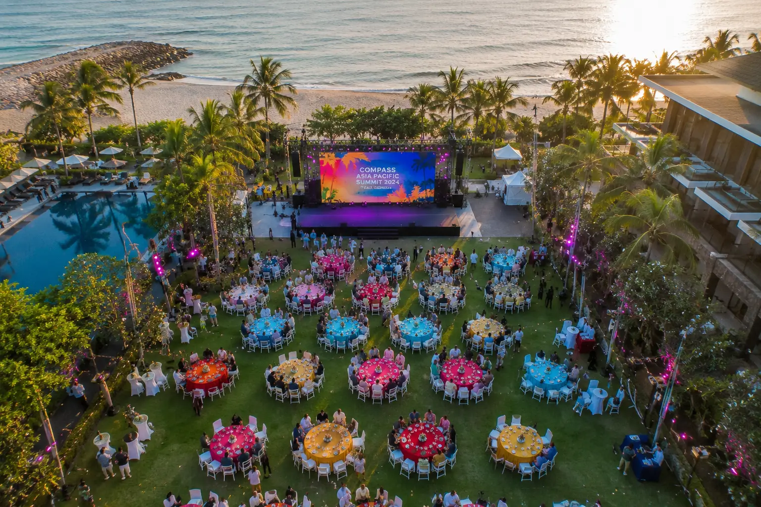 Professional drone videography capturing a luxury corporate event in Bali with colorful event branding, oceanfront venue, tropical resort setting, and aerial event coverage.